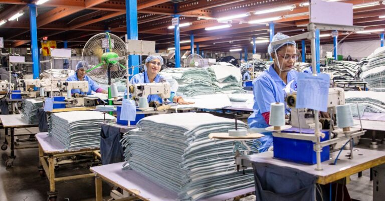 Công nhân nữ làm việc tại dây chuyền may mặc trong nhà máy, xung quanh là máy móc và sản phẩm dệt may.
